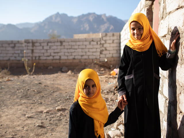 Two young girls, wearing matching headscarves, hold hands by a wall in Yemen.