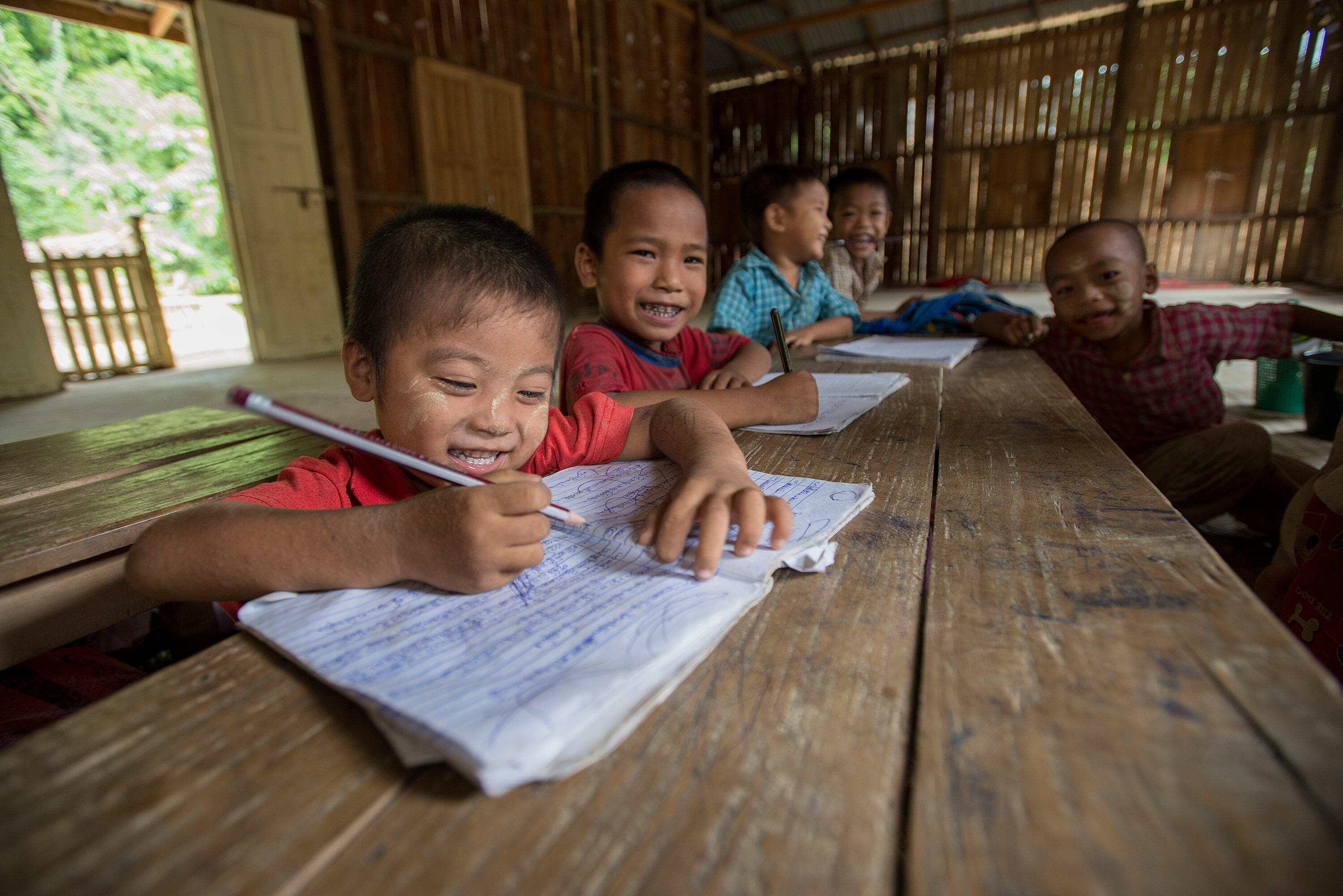 Smiling young boys in Myanmar write in their notebooks at a wooden table in tehir bamboo classroom.