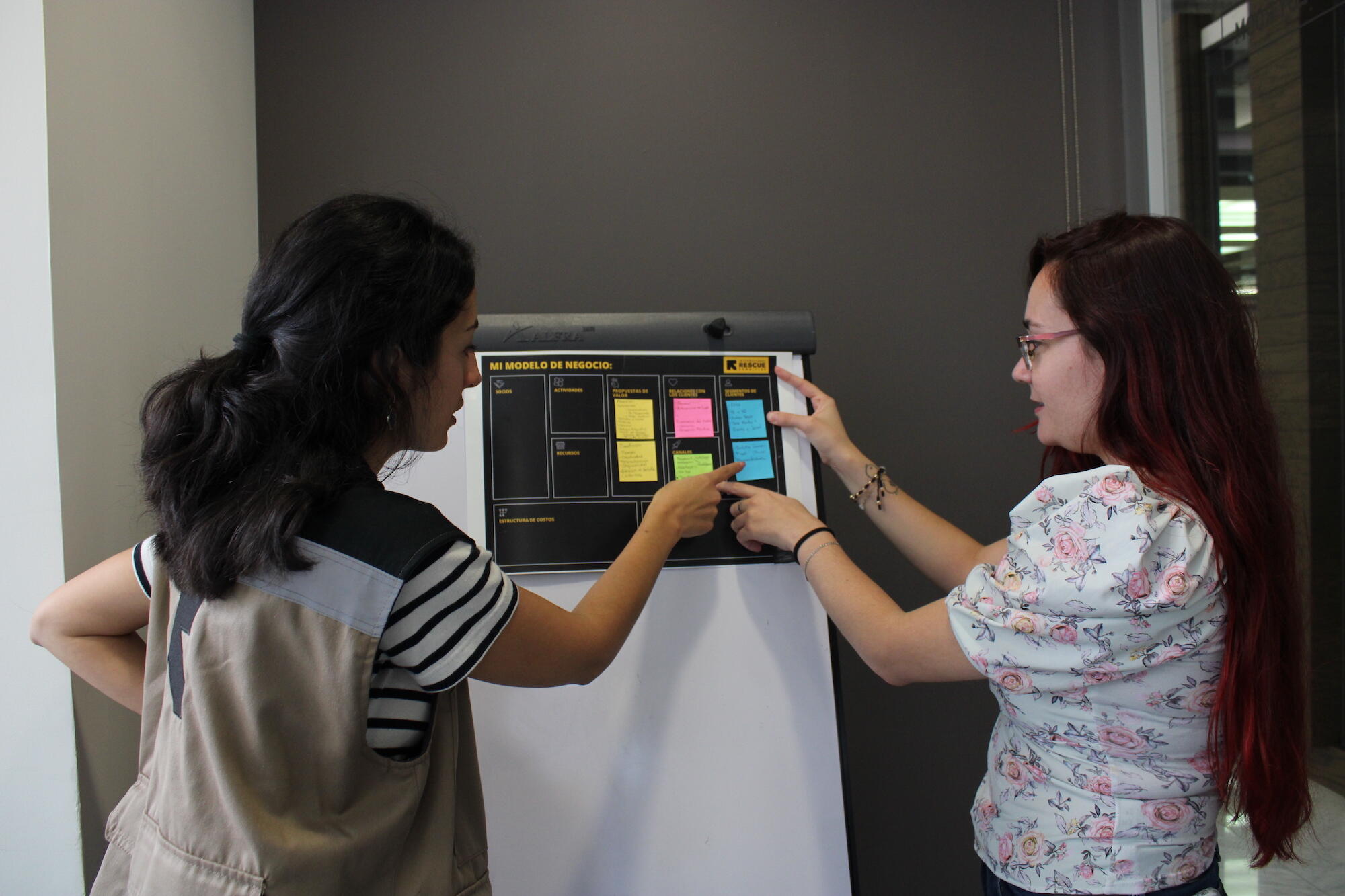 Two women stand in front of a white board with colored sticky notes.