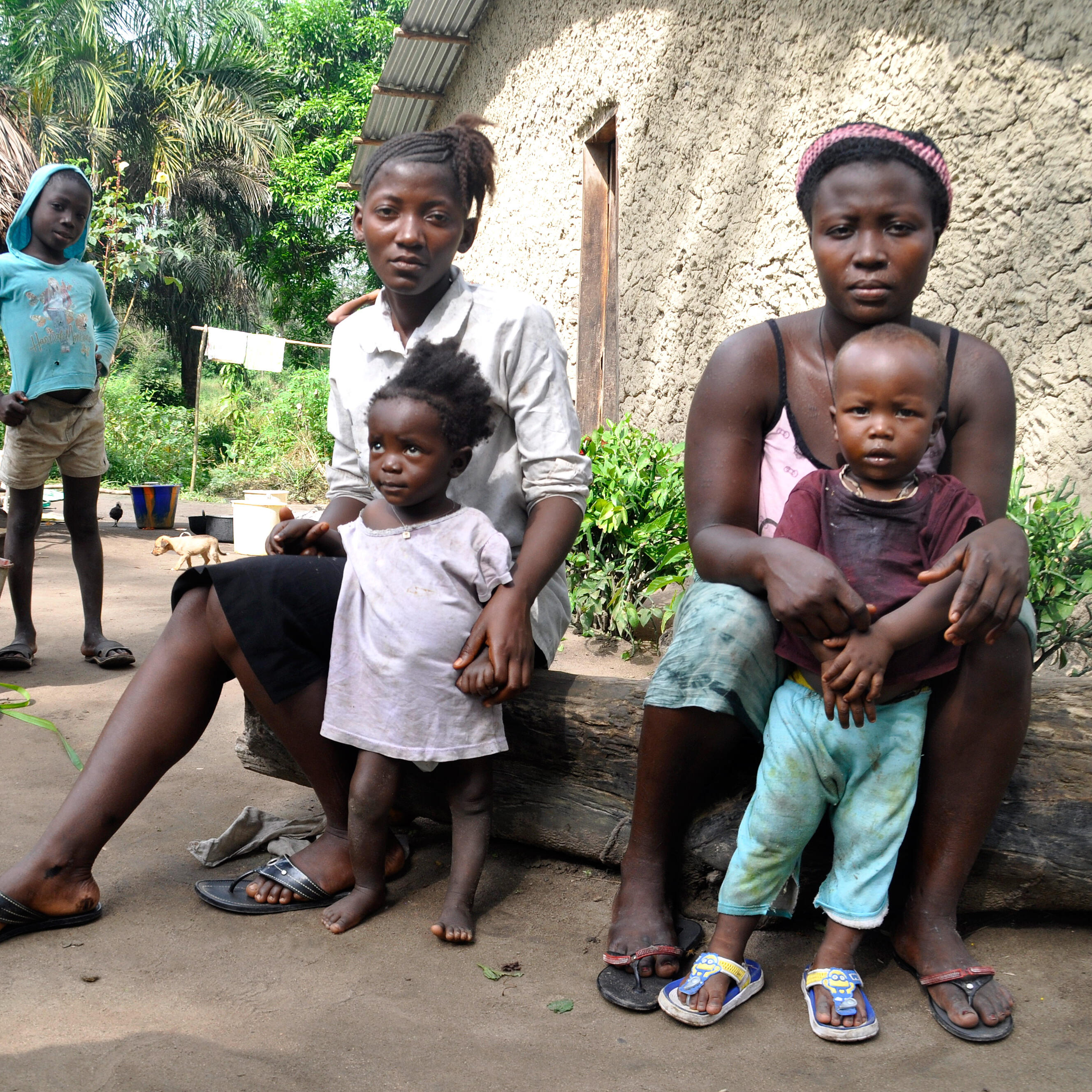 Two women sit on a bench and hold their young children, while a man and a girl stand in the background.
