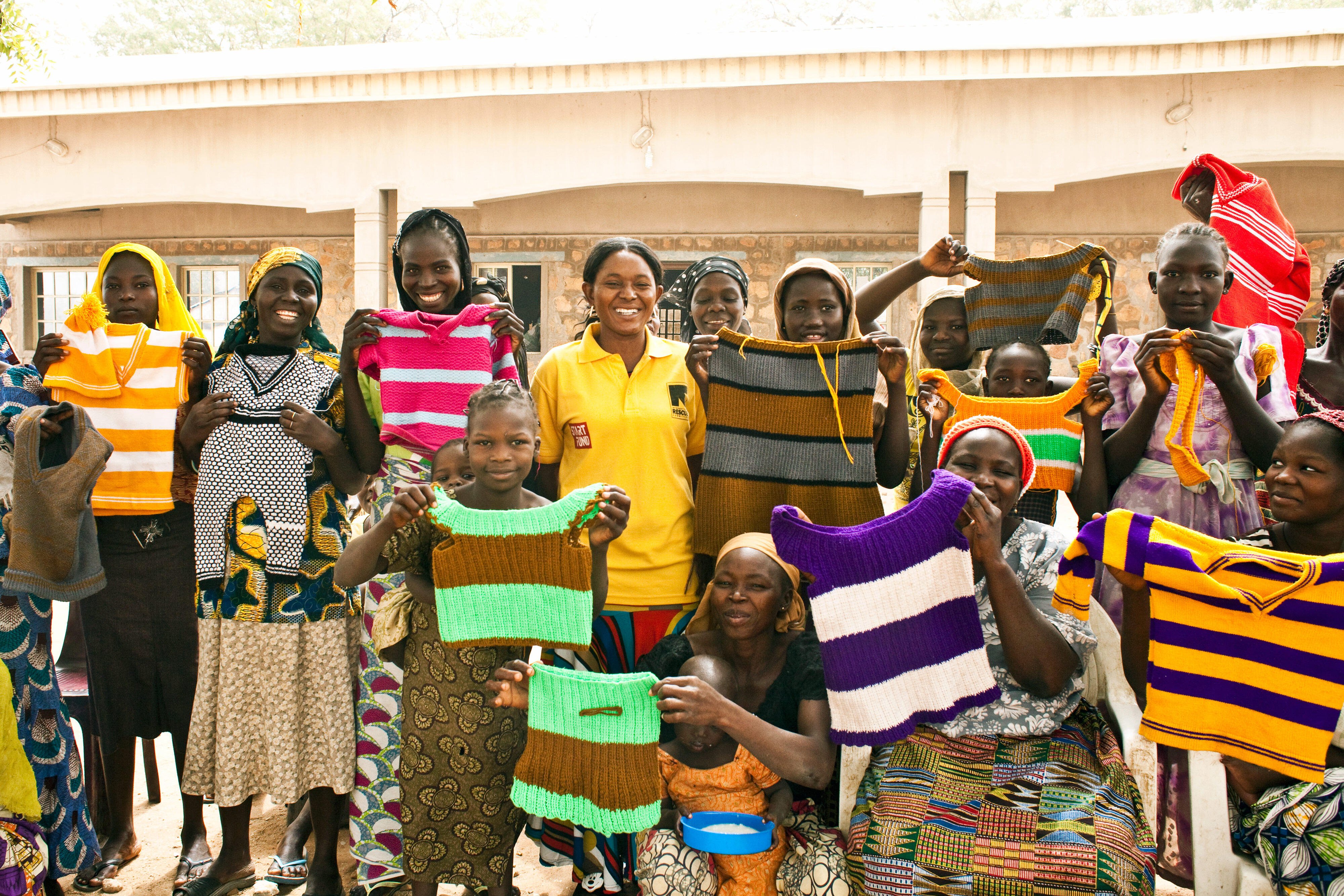 A group of women who are part of an IRC livelihoods program in Nigeria stand outside, lifting hand-knitted tops to show them off.  