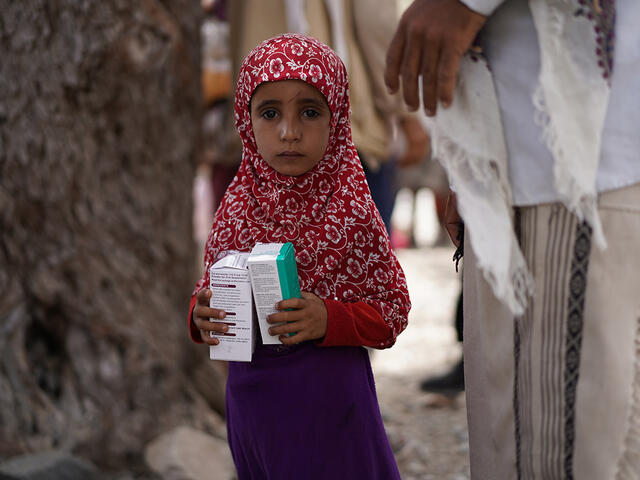 A three year old girl in a pink headscarf holding two boxes of medicine