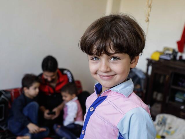 Five-year-old Murad smiles at the camera with his mum in the background making masks in Syria