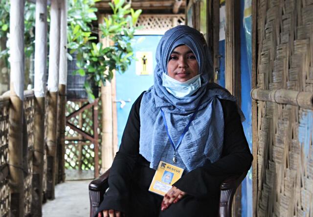 Yasmin Ara sits on a chair on a porch, looking at the camera. She is wearing a blue head scarf, black jacket and IRC lanyard.