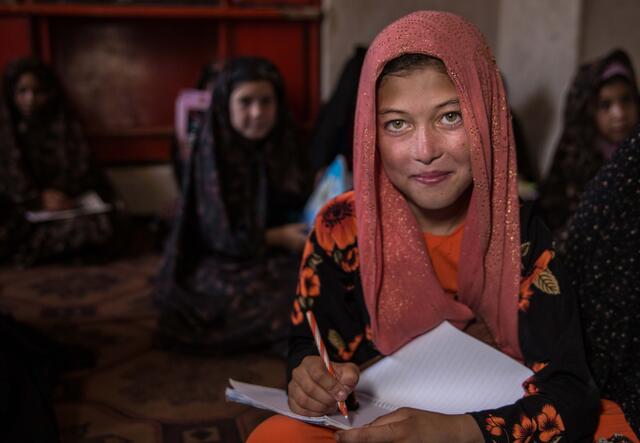 A young girl in the classroom poses for a photo while writing in her notebook.