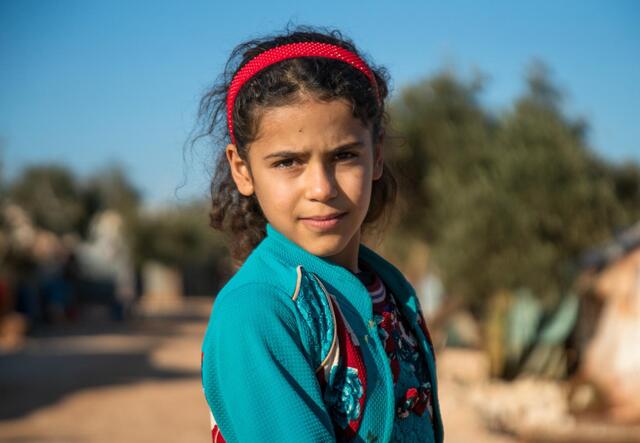 10-year-old Heba looks at the camera. She wears a red head band and green cardigan.